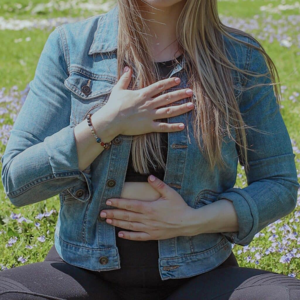 Jillian, travel and wellness influencer, sitting cross-legged on a grassy field, engaging in a mindful meditation practice. Her hands are positioned on her chest and abdomen, expressing a sense of peace and self-connection against a serene outdoor background.