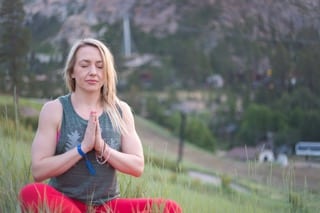 Jillian, travel and wellness influencer meditating outdoors with her hands in a prayer position, eyes closed, and a gentle smile on her face. The background shows a soft-focus hillside, enhancing the tranquil setting of her meditation practice.