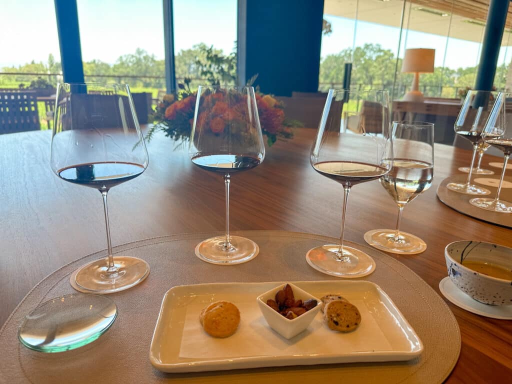 A wine tasting setup at Opus One winery in Napa Valley, featuring three glasses of red wine, a glass of water, and a small plate with assorted snacks including nuts and savory biscuits. The scene is set against large windows showcasing the vineyard outside, providing a serene and elegant ambiance for the tasting experience.