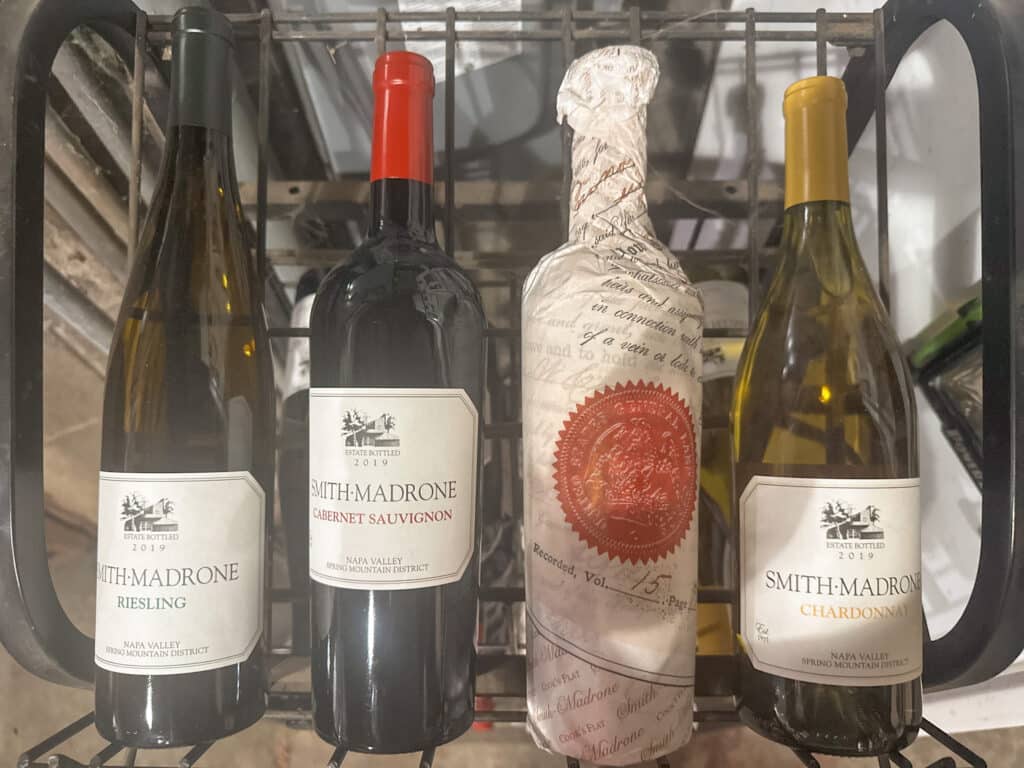 A selection of Smith-Madrone wines from Napa Valley, including bottles of Riesling, Cabernet Sauvignon, and Chardonnay, is displayed on a metal rack. One bottle is wrapped in decorative paper, showcasing the winery's branding and attention to detail, highlighting the variety and quality of their estate-bottled wines.