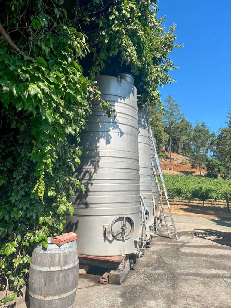 Large stainless steel fermentation tanks at Smith Madrone winery in Napa Valley are partially shaded by overhanging greenery. A wooden wine barrel and a ladder are visible nearby, set against a backdrop of vineyards and trees under a clear blue sky, illustrating the winemaking process in this picturesque vineyard.