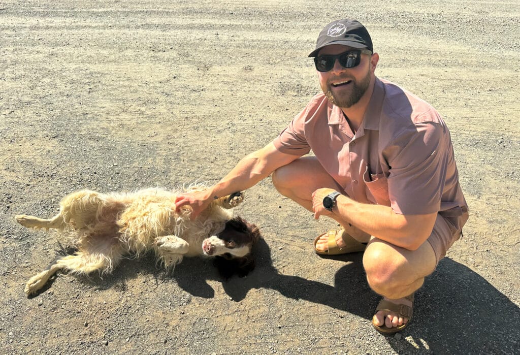 Brandon kneels on a gravel path, smiling and wearing sunglasses and a cap, as he pets Tucker, the friendly winery dog at Smith Madrone. Tucker, a fluffy dog with a white and brown coat, lies on his back enjoying a belly rub under the warm sun.