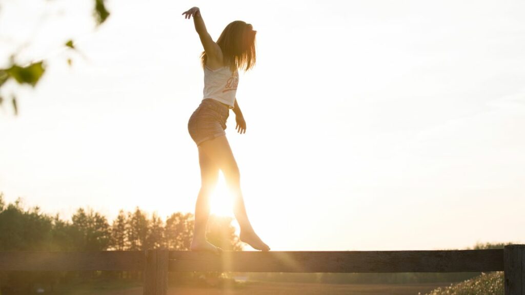 The image shows a woman balancing on a wooden beam with one arm raised as the sun sets behind her. The sunlight creates a bright, glowing silhouette around her, and the scene has a peaceful, serene feel with a natural landscape in the background. The warm lighting emphasizes the calmness and freedom of the moment.