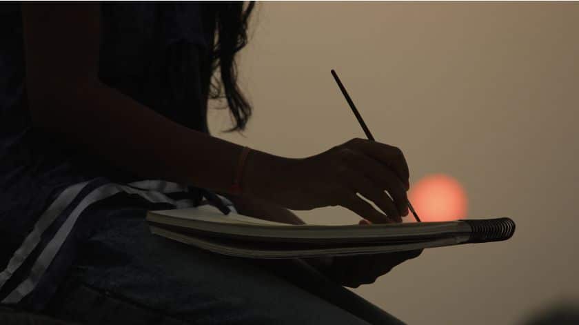 The image shows a close-up silhouette of a person sketching or painting on a notepad during sunset. The warm glow of the setting sun is visible in the background, casting a serene ambiance over the scene. The focus is on the person's hand holding a brush or pencil, engaged in a peaceful and creative moment.