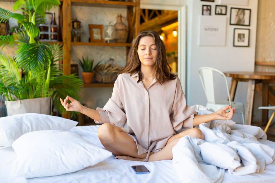 The image shows a woman sitting cross-legged on a bed, practicing meditation with her eyes closed and hands resting on her knees in a peaceful pose. She is wearing a casual, comfortable outfit in a cozy, boho-styled room with plants and wooden decor in the background. A phone is placed beside her on the bed, symbolizing a break from technology, contributing to a sense of calm and digital detox.